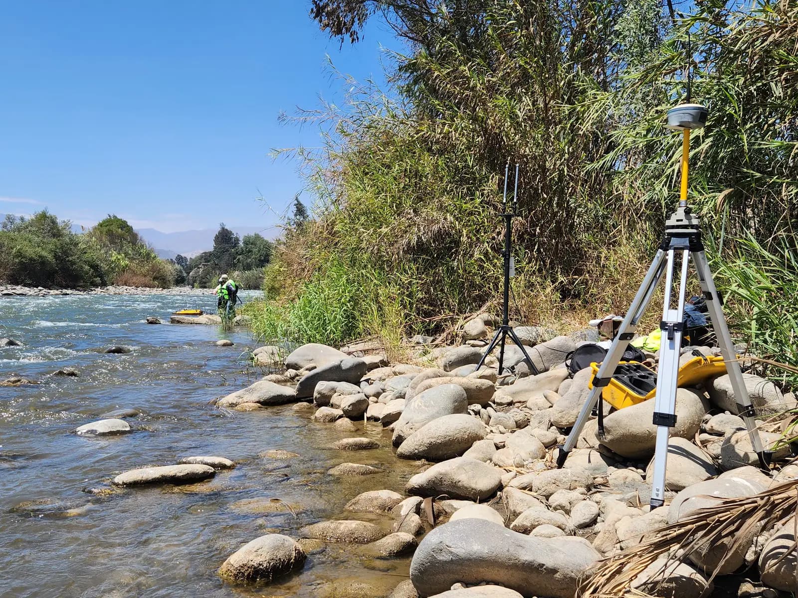 Base GNSS en la ribera de un río mientras la brigada trabaja en el cauce
